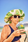 Woman with strawberry hat and sunglasses, holding a drink at the beach.